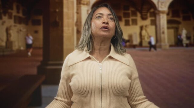 Woman mouth open looking up inside historic building with statues and arches, arms slightly raised; reverence contemplation.