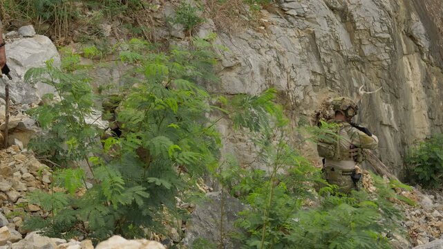 Special forces soldiers, including a ghillie-suited sniper, secure a tactical overwatch position during a reconnaissance mission in a rugged, rocky terrain.