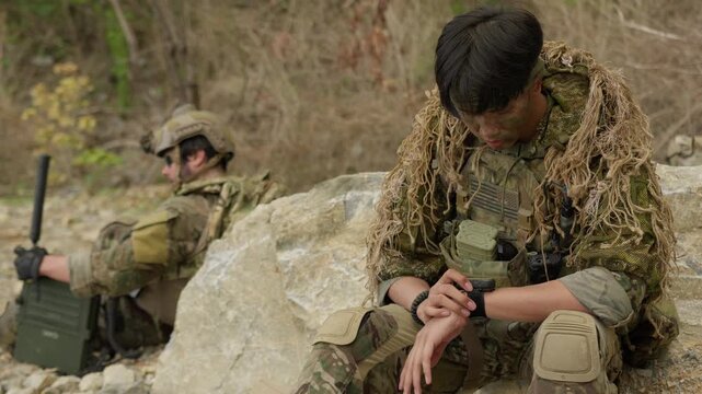 A special forces sniper in a ghillie suit synchronizes his tactical watch for a covert operation, with a radioman coordinating in the rocky wilderness.