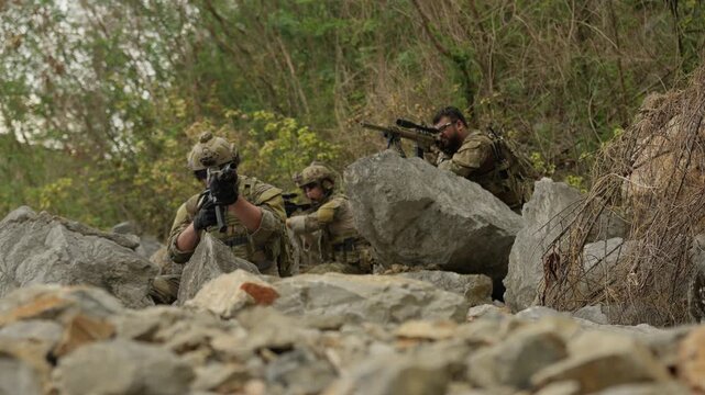 A military squad establishes a tactical position behind rocks, the sniper providing overwatch during a covert special operations mission in rugged terrain.
