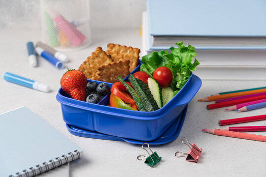 Blue school lunch box with berries crackers and vegetables on light background, side view with stationery, back to school concept