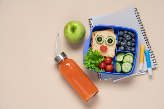 Kids school lunch box with fun face sandwich vegetables and blueberries in blue container and carrot juice on light beige background, back to school concept