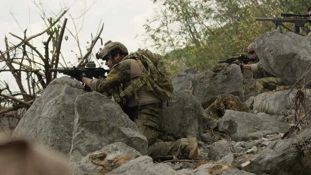 Special forces soldiers in full combat gear take defensive positions behind rocks, aiming their rifles during a tactical reconnaissance mission in hostile territory.