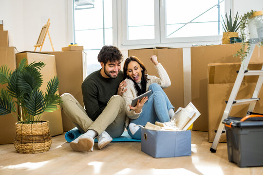 A happy young couple sitting on the floor among moving boxes and celebrating while looking at a digital tablet