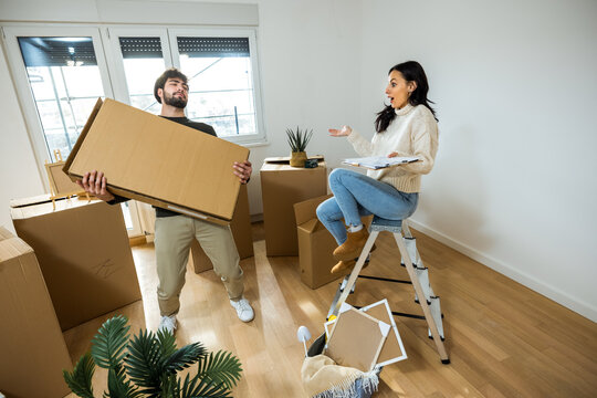A young woman holding a checklist on a stepladder while a man struggles to lift a heavy cardboard box