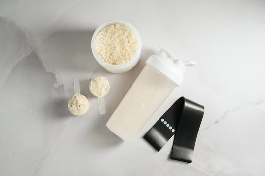 Protein shake, whey powder and resistance band on white background, fitness flat lay