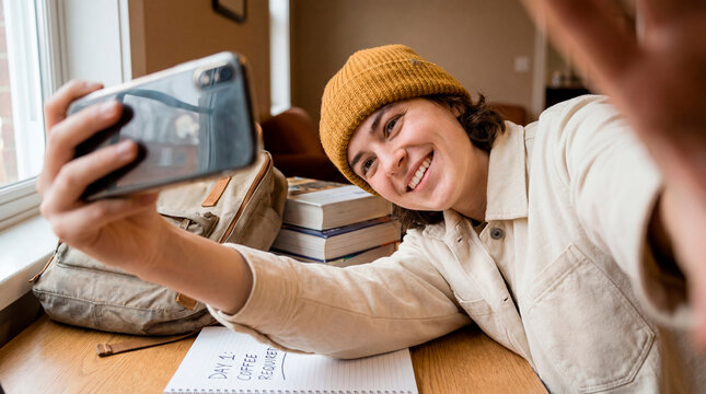 Playful young woman in beanie taking selfie with smartphone among study books