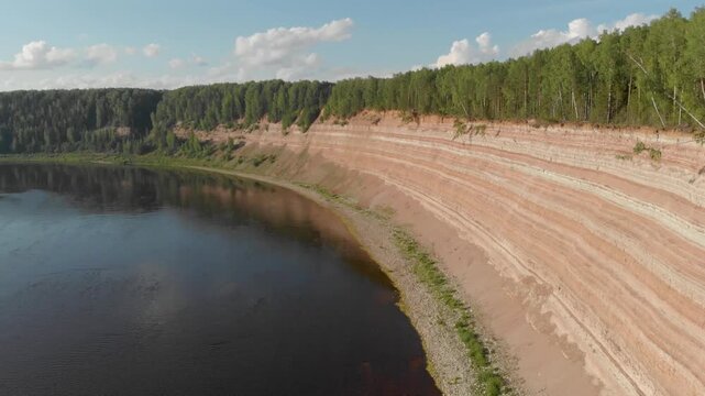Aerial view shows a calm river beside a steep red clay cliff. Trees line the cliff's top under a bright blue sky. Soft clouds drift above the tranquil water surface