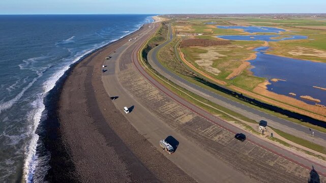 Aerial view of a curved coastal dike with a road, vehicles, ocean waves, parallel inland roads, green fields, ponds, and wetlands under clear daylight.