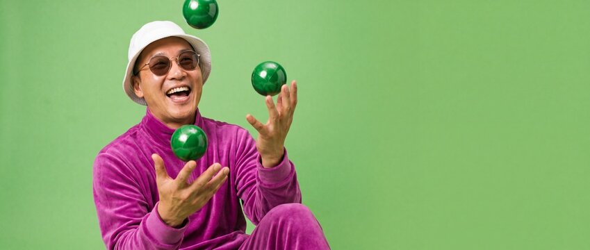 Happy Asian man juggling green balls wearing purple velour tracksuit and white bucket hat, cheerful mature guy playing against solid green copy space background, fun lifestyle banner concept.