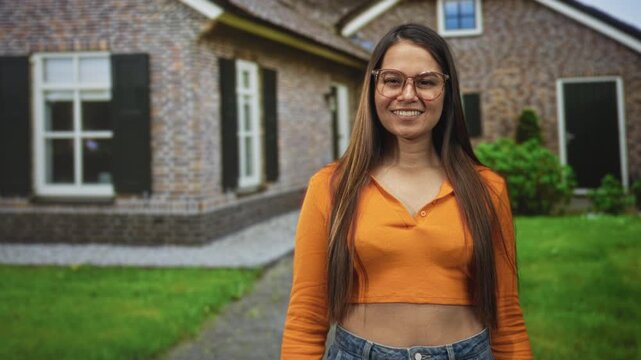 Woman smiling with visible midriff in orange crop top and denim jeans, wearing glasses, standing on a lawn pathway in front of a brick building; happiness casual confidence.