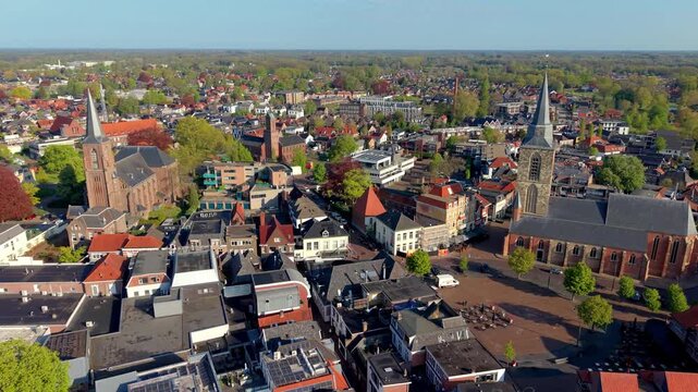 Aerial view of a European town with historic churches, dense buildings, red‑tiled roofs, streets, greenery, and surrounding residential areas under clear daylight.