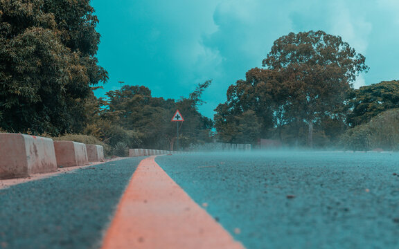 Misty wet paved road leading toward a triangular sign under a thick cloudy sky surrounded by large trees.