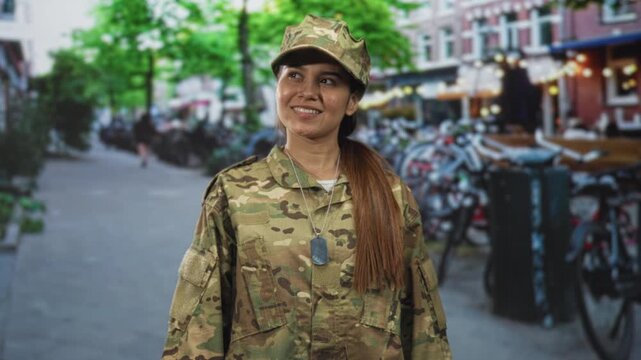 Woman soldier in camouflage uniform smiling with dogtag visible, standing on street lined with bicycles and cafes; pride duty.
