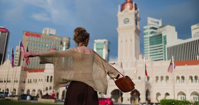 A happy young woman with a joyful smile turns and poses gracefully against the majestic backdrop of the historic Sultan Abdul Samad Building in vibrant Kuala Lumpur, capturing the spirit of travel and