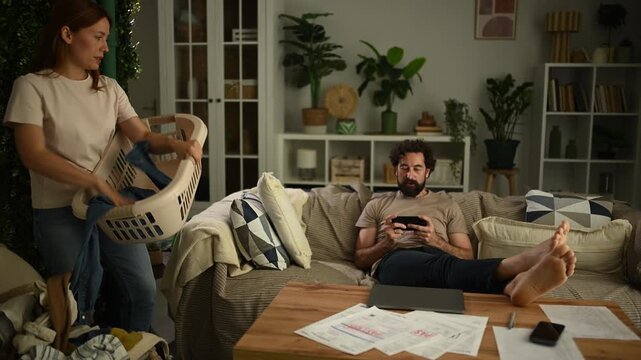 Angry woman throwing laundry at lazy husband playing video games on the couch, symbolizing unequal domestic labor