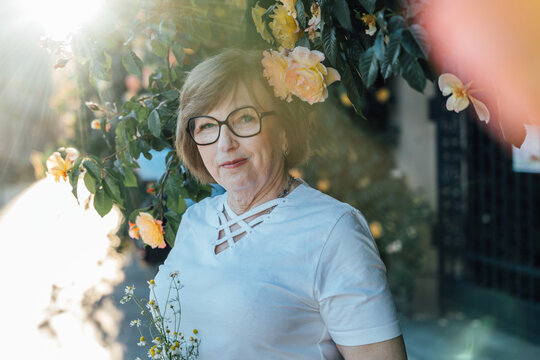 Elegant Senior Woman Standing Near Blooming Rose Wall In Warm Summer Evening Light