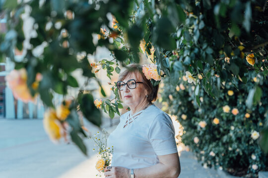 Thoughtful Senior Woman Holding Chamomile Bouquet Beneath Blooming Yellow Rose Garden Arch