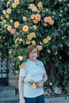 Elegant Senior Woman Standing Near Abundant Yellow Rose Wall With Summer Flowers