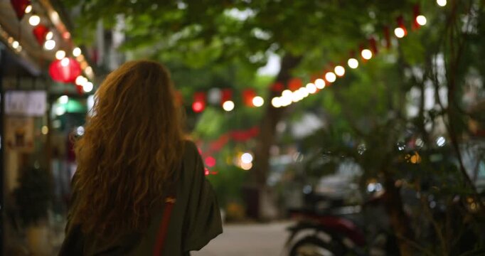 A young woman enjoys a phone conversation while walking at night on the vibrant Train Street in Hanoi, Vietnam, surrounded by charming string lights and red lanterns, experiencing the city's lively