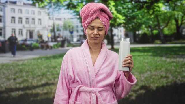 Woman holding shampoo bottle, pink bathrobe and towel turban, face visible, standing in park; annoyance.