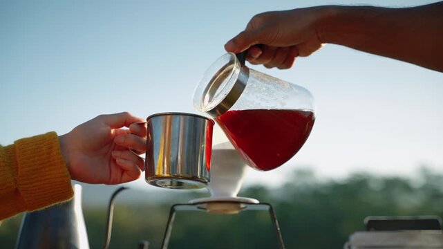 Close-up of hands pouring freshly brewed coffee from a glass carafe into a stainless steel camping mug beside a pour-over dripper outdoors. Warm morning light, camping lifestyle, relaxation, and natur