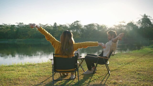 Rear view of a couple sitting in camping chairs beside a lake and stretching in the morning sunlight. Outdoor recreation, relaxation, wellness, travel, and peaceful nature lifestyle concept.
