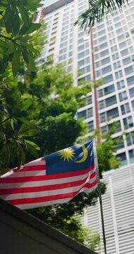 The Malaysian flag, 'Jalur Gemilang', proudly waves against a modern city skyscraper, symbolizing national pride and identity amidst green urban foliage.