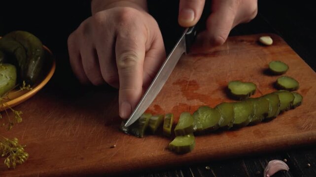 Hands of a person slicing pickle on a wooden cutting board with a bowl of whole pickles and garlic cloves visible in the background