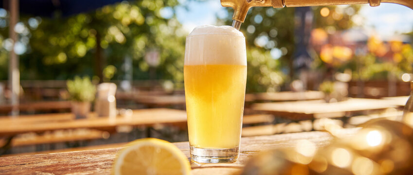 Cold draft beer in pint glass on wooden table at sunny outdoor beer garden
