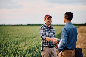 Fototapeta premium Happy mature farmer handshaking with insurance representative while standing in field.