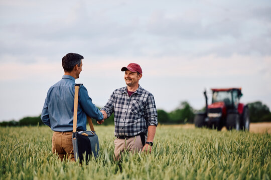 Happy agronomist shaking hands with his insurance consultant while standing in wheat field.