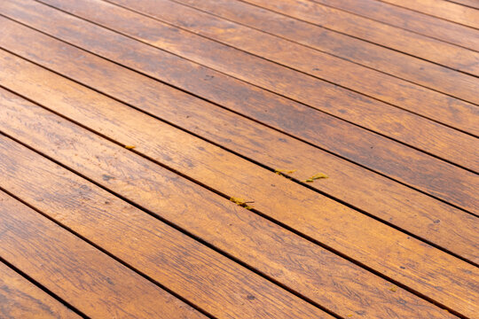 Detail of a wooden surface with parallel planks at a diagonal angle. Shows natural grains, reddish tones, and small organic debris. Ideal for design and architecture backgrounds.