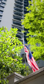 The National Flag of Malaysia, the Jalur Gemilang, Gracefully Flutters in the Gentle Breeze Against a Bright Backdrop of a Modern High-Rise Building and Lush Green Trees