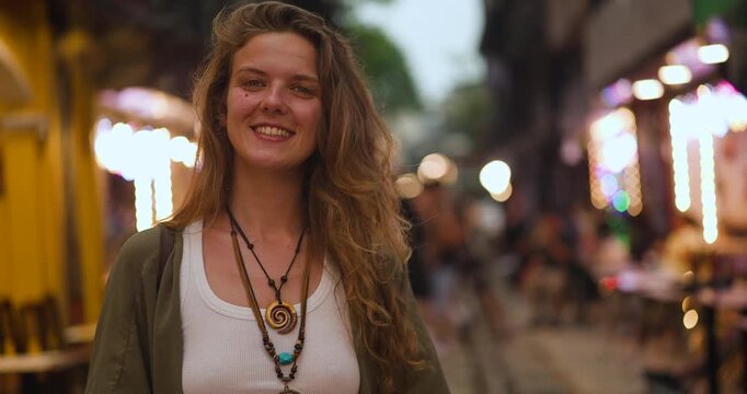 Happy young woman captures memories with her smartphone at the bustling Train Street in Hanoi, Vietnam, smiling brightly at the camera amidst the vibrant evening lights of the city.