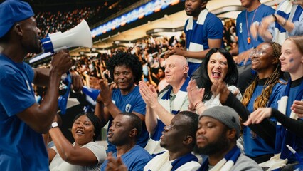 Excited football fan leader using bullhorn to unite blue supporter group in stadium stands during live soccer match atmosphere, passionate football fans in the supporter section singing chants. © DC Studio