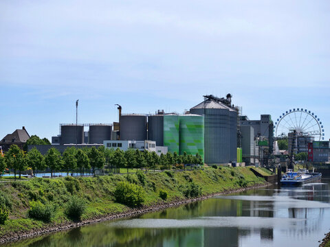 A wide landscape shot captures an active 
industrial port and canal. The riverbank is 
heavily vegetated with green bushes and 
trees. Large grey, green, and silver industrial 
storage tanks and grain