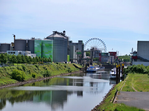A scenic shot of an active industrial canal and 
river port. The water in the canal reflects the 
industrial complex, storage tanks, and large 
Ferris wheel standing prominently in the 
background. Al