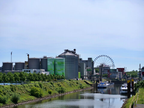In the  foreground, a tranquil river canal reflects a 
green, grassy embankment. Across the water 
stands a large industrial facility with distinct 
light green and grey painted silos, various 
wareho