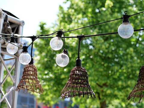 : A close-up, selective focus 
shot of outdoor string lights hanging at a 
venue or caf&eacute;. The garland features 
round glass light bulbs interspersed with 
decorative, bell-shaped woven wicker 
lampsha