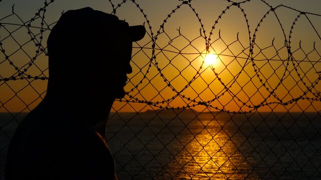 Prisoner looking at sunset through barbed wire fence. Prisoner silhouette gazing through barbed wire fence, embodying deep loneliness and yearning for freedom against dramatic sunset landscape