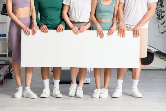 Diverse group of fitness enthusiasts holding blank white banner in modern gym, copy space for health and wellness message