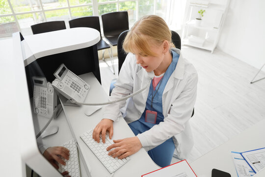 Medical receptionist answering phone call and typing on computer at hospital front desk, the professional woman in a lab coat and scrubs is providing patient service and support