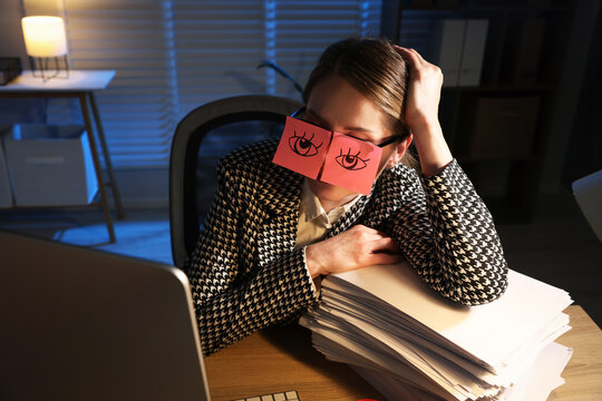 Workaholic. Woman in glasses and fake eyes drawn on sticky notes with stack of documents and computer at wooden desk indoors at night