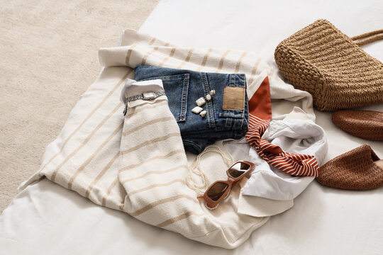 Summer vacation travel outfit flat lay with jeans, striped sweater, straw bag, and accessories on bed