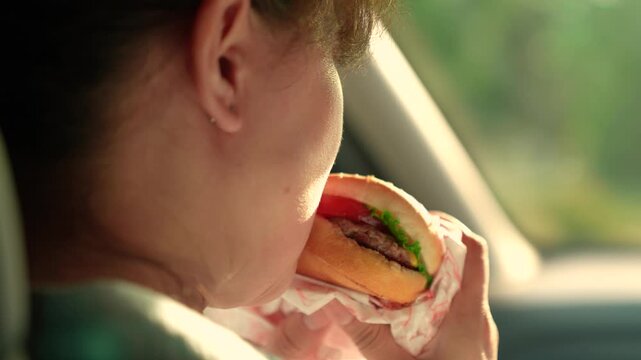 Young woman satisfies hunger during car ride. Happy woman snack in car. Girl eats burger car. Hungry girl eats fast food burger car. Fast food automobile. Snack road. Delicious food satisfies hunger
