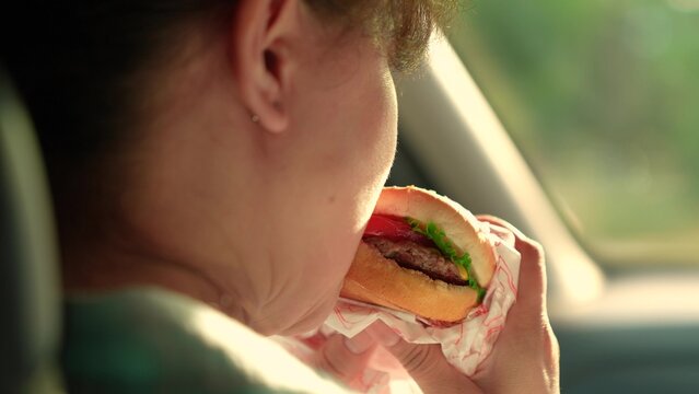 Young woman satisfies hunger during car ride. Happy woman snack in car. Girl eats burger car. Hungry girl eats fast food burger car. Fast food automobile. Snack road. Delicious food satisfies hunger