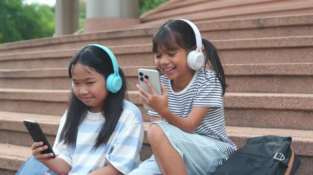 Cute young Asian school girls listening music via wireless headphones and digital tablet on campus step. Happy primary classmates enjoying e-learning back to school study loop .