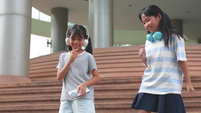 Cute young Asian school girls listening music via wireless headphones and digital tablet on campus step. Happy primary classmates enjoying e-learning back to school study loop  clip.