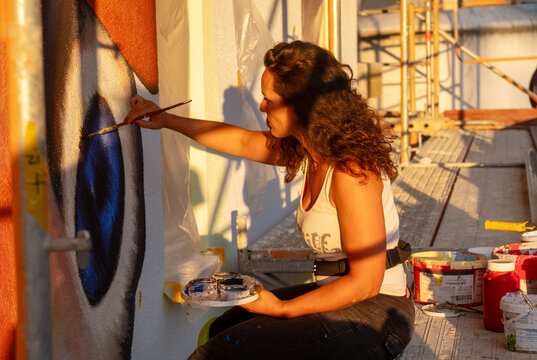 Female mural artist paints a large facade artwork from scaffolding during golden hour. Warm evening sunlight highlights creativity, craftsmanship and the process of architectural art